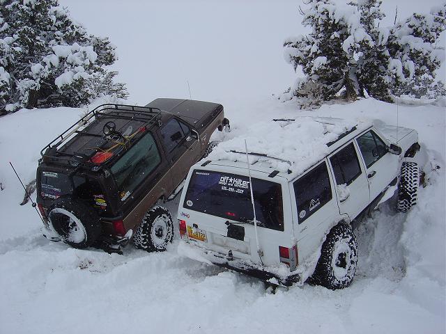 Brown SUV and white Jeep Cherokee stuck in deep snow, trees covered in snow in background