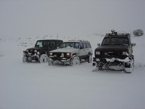 1986 Ford Bronco II - Three SUVs are parked in a snowy landscape, with one black Jeep Wrangler, a white Jeep Cherokee, and a dark SUV partially covered in snow.