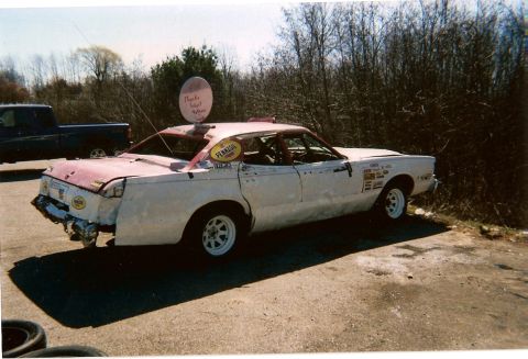 1977 Mercury Cougar - This heavily modified sedan, likely prepared for demolition derby, is parked outdoors next to a dark pickup truck.