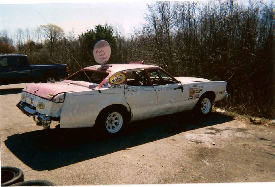 Modified sedan with racing decals and a round sign on top, parked next to a pickup truck