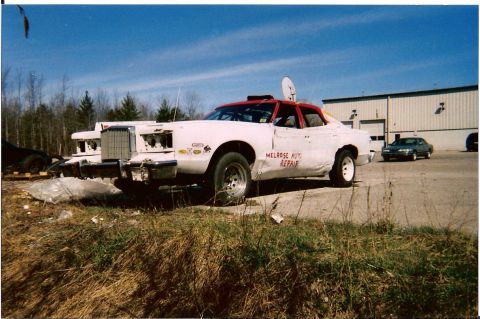 1977 Mercury Cougar - A white race car with a red roof and visible damage is parked in front of a commercial building.