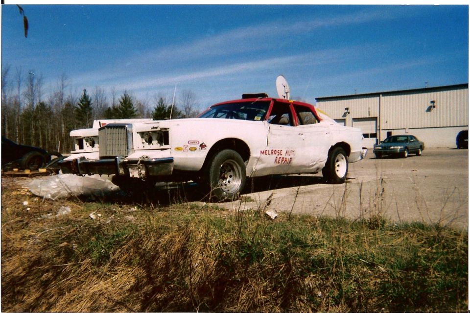 White race car with red roof, dented body, and "MELROSE AUTO REPAIR" text, parked outside a building