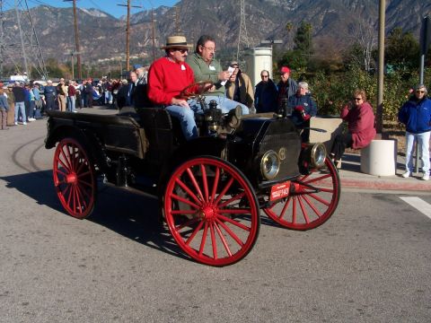 1990 Ford Bronco - Two men ride in an early 20th-century touring car with large red spoked wheels, observed by onlookers.