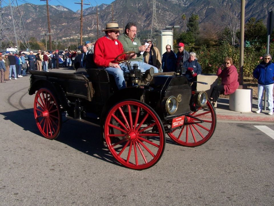 Antique black touring car with red wheels, two men in front, crowd in background