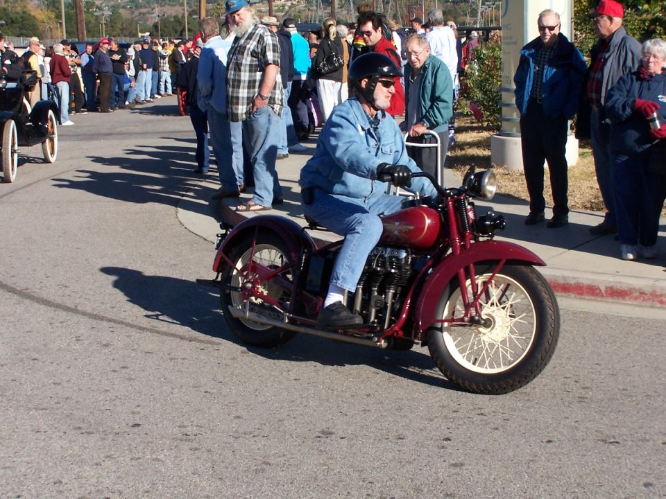 Man in denim jacket riding a red vintage motorcycle with a crowd watching