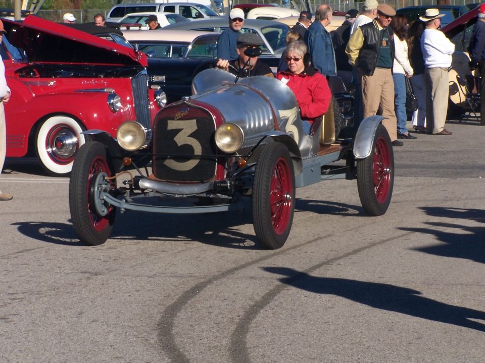 Two people in a silver vintage race car with the number 3 on the front. Red wheels and red accents