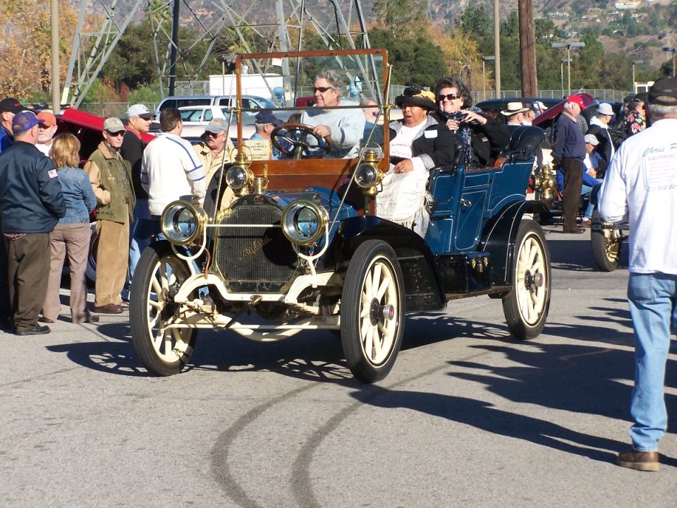Blue antique touring car with people inside, driving on a paved road with spectators around