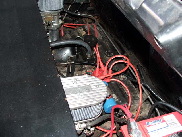 Carbureted rear engine bay with finned cover, red plug wires, and a battery