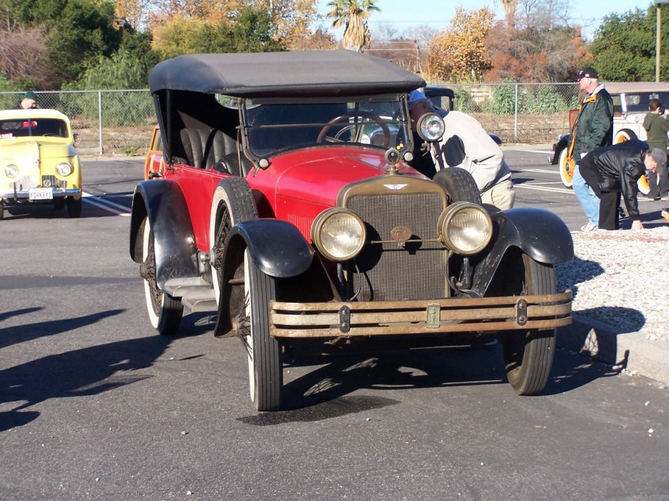 Red touring car with black fenders and a black soft top, parked outdoors. Yellow car visible in background