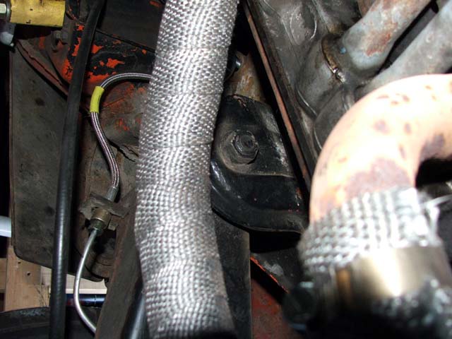 Braided heat-shield-wrapped hose beside rusty exhaust pipe in vehicle undercarriage