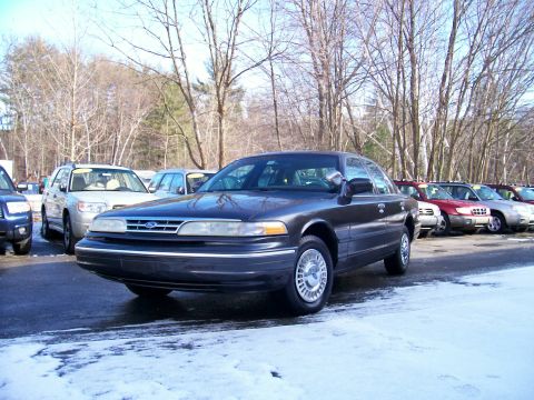 2001 Ford Crown Victoria - A dark sedan is parked in a lot with other vehicles visible in the background, under a bright sky.