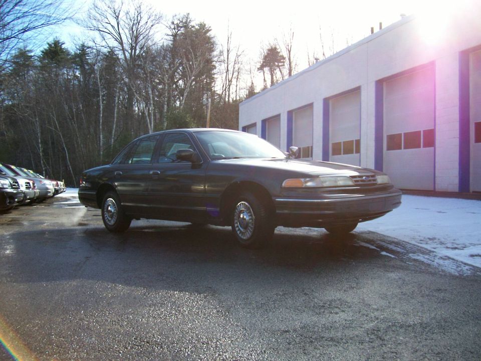 Dark 2001 Ford Crown Victoria sedan parked on wet asphalt, front quarter view, with a building and trees in the background