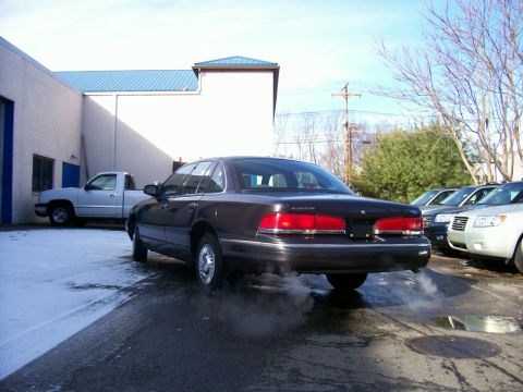 2001 Ford Crown Victoria - A dark sedan is seen from the rear with exhaust fumes visible, parked in a lot with other vehicles.