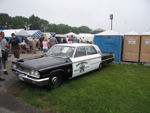 1991 Ford LTD Crown Victoria - black-and-white vintage sedan lettered Mayberry Sheriff's Office sits at a crowded outdoor car show near tents and portable toilets.