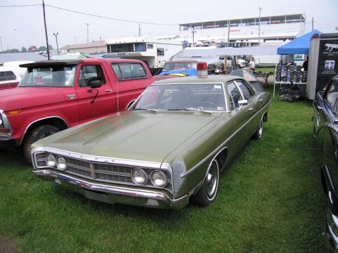 1991 Ford LTD Crown Victoria - green vintage Ford four-door sedan with FORD hood lettering and a roof beacon sits on grass beside a red pickup at an outdoor car show.