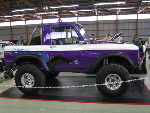 1991 Ford LTD Crown Victoria - Side profile of a lifted purple-and-white early Ford Bronco with oversized tires, blue flame graphics and Bronco script, on an indoor displ…