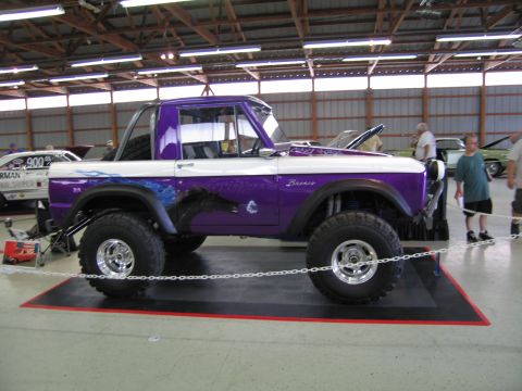 1991 Ford LTD Crown Victoria - Side view of a lifted purple-and-white early Ford Bronco with big tires and Bronco fender script, displayed indoors as spectators walk past.