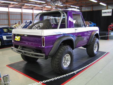 1991 Ford LTD Crown Victoria - Rear view of a lifted purple-and-white early Ford Bronco with roll bar and spare tire, parked behind a chain barrier at an indoor show.