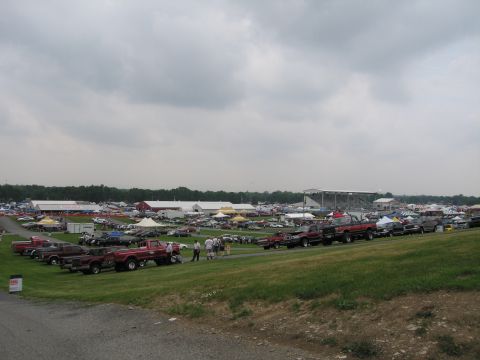 1991 Ford LTD Crown Victoria - panoramic view over a crowded outdoor car show and swap meet with rows of vehicles, tents and grandstands beneath an overcast sky.
