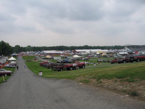 1991 Ford LTD Crown Victoria - Rows of pickups, cars and vendor tents fill a large grassy car-show field under a gray cloudy sky, with a paved walkway in the foreground.