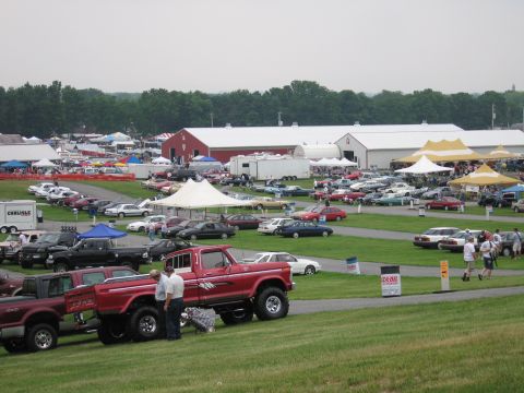 1991 Ford LTD Crown Victoria - red lifted pickup and rows of vehicles and tents fill a busy outdoor car-show field, with white show buildings in the background.