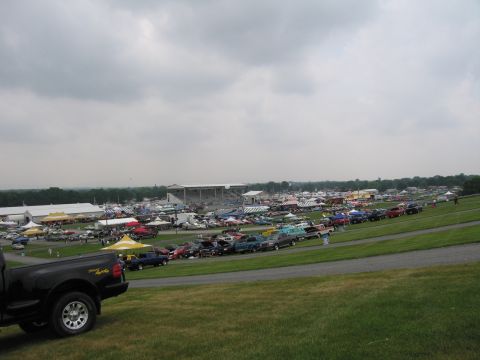 1991 Ford LTD Crown Victoria - View across a packed outdoor car-show field of cars, tents and grandstands under a cloudy sky, with a black pickup tailgate in the foregrou…