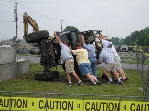 1991 Ford LTD Crown Victoria - Several men in shorts work together to tip a small dark truck onto its side on grass, with caution tape and an excavator in the background.