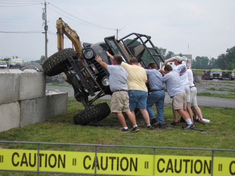 1991 Ford LTD Crown Victoria - Several men work together to right an off-road tube-frame buggy tipped against concrete barrier blocks, behind yellow CAUTION tape at an ou…