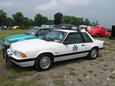 1991 Ford LTD Crown Victoria - white fox-body Ford Mustang police interceptor with a sheriff-style star door emblem and roof light bar, parked on grass among other show c…