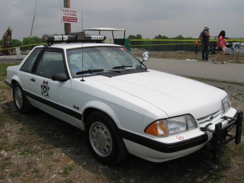 1991 Ford LTD Crown Victoria - white fox-body Ford Mustang police car with a front push bar, roof light bar and star door emblem sits on gravel at an outdoor show.