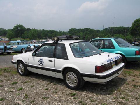 1991 Ford LTD Crown Victoria - Rear three-quarter view of a white fox-body Ford Mustang police car with a star door emblem and roof light bar, parked among classic cars o…