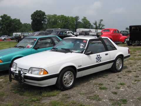 1991 Ford LTD Crown Victoria - white fox-body Ford Mustang police car with front push bar and star door emblem parked beside a teal Mustang on grass at an outdoor show.