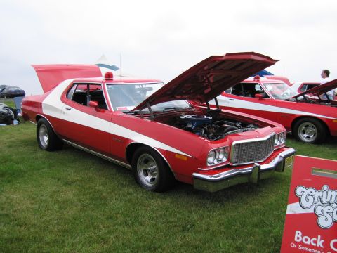 1991 Ford LTD Crown Victoria - red Ford Gran Torino with a white vector stripe and open hood, displayed on grass at an outdoor car show beside a matching car.