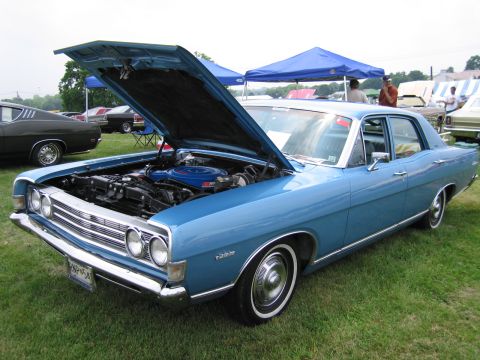1991 Ford LTD Crown Victoria - light blue 1960s Ford four-door sedan with open hood, chrome grille and full wheel covers parked on grass under tents at a car show.