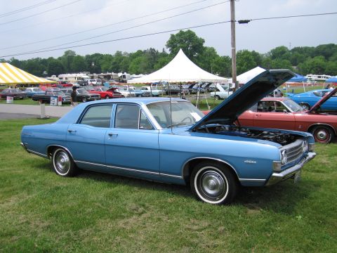 1991 Ford LTD Crown Victoria - Side profile of a light blue 1960s Ford four-door sedan with hood raised, parked on grass with a large white tent behind it at a car show.