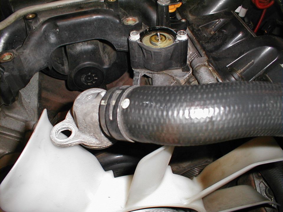 Close-up of engine thermostat housing with a large hose and fan visible below. Supermotors.net logo in lower right