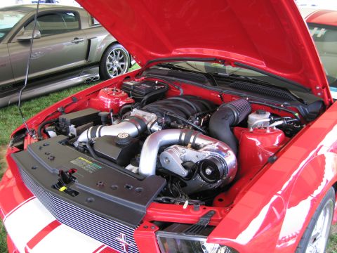 1991 Ford LTD Crown Victoria - engine bay of a red Ford Mustang showing a polished supercharger and detailed components under a raised hood at an outdoor show.