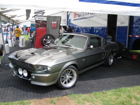 1991 Ford LTD Crown Victoria - gray Eleanor-style Shelby Mustang fastback with white stripes and dual driving lights displayed under a vendor tent on flooring at a show.