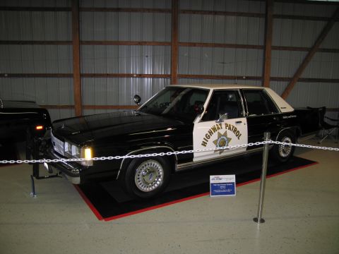 1991 Ford LTD Crown Victoria - black and white CHP Highway Patrol Ford sedan with gold star decals sits roped off inside a metal exhibition building.
