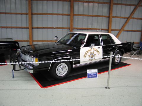 1991 Ford LTD Crown Victoria - Front three-quarter view of a black and white Highway Patrol Ford sedan with a gold star door shield, displayed behind a chain barrier.