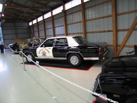 1991 Ford LTD Crown Victoria - black and white Highway Patrol sedan among a row of police cars displayed inside a metal exhibition building.