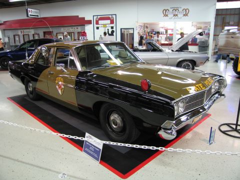 1991 Ford LTD Crown Victoria - tan and black vintage State Police Ford sedan with a single red roof light, displayed on a red mat inside an indoor exhibit.