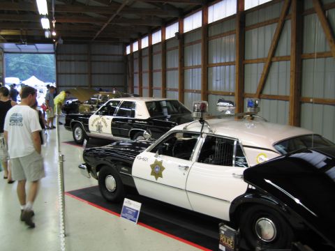 1991 Ford LTD Crown Victoria - Several black and white police cruisers with roof light bars and push bars line up inside a metal exhibition barn as visitors look on.