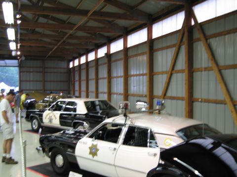 1991 Ford LTD Crown Victoria - Black and white police cruisers fitted with roof light bars and spotlights are lined up along the wall of a metal exhibition hall.