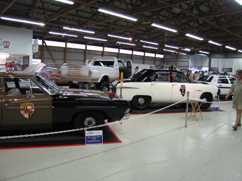 1991 Ford LTD Crown Victoria - tan State Police car sits beside black and white cruisers and a pickup truck inside a brightly lit exhibition building.