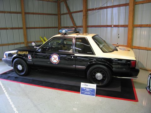 1991 Ford LTD Crown Victoria - Black-and-tan Florida State Trooper Ford Mustang 5.0 notchback patrol car with lightbar, displayed indoors on a stand.