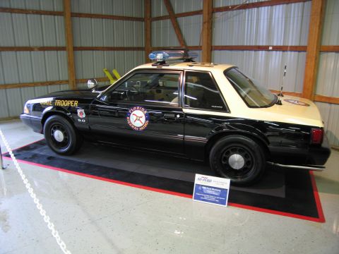 1991 Ford LTD Crown Victoria - Black-and-tan Florida Highway Patrol Mustang 5.0 patrol car with rooftop lights and info placard in an indoor exhibit.