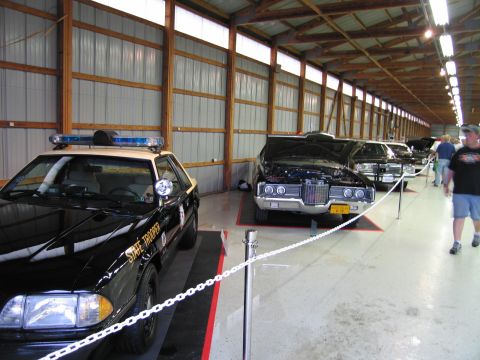 1991 Ford LTD Crown Victoria - Indoor display row with a black-and-tan State Trooper patrol car in front and classic cars behind a chain barrier.