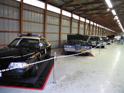 1991 Ford LTD Crown Victoria - Black State Trooper patrol car at the head of a chained-off row of classic vehicles inside a metal exhibit building.