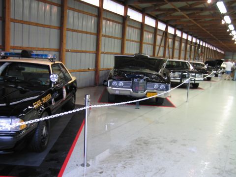 1991 Ford LTD Crown Victoria - Row of display cars behind chain stanchions, led by a State Trooper patrol car beside a chrome-grilled classic coupe.